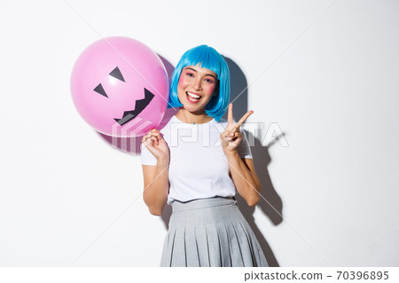 Image of happy asian girl celebrating halloween in blue anime wig, holding pink balloon with scary face and showing peace gesture 70396895