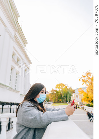Portrait of a beautiful adult young woman on the background of autumn in the Park in a medical face mask with a phone in her hands Portrait of a beautiful adult young woman on the background of autumn in the Park in a medical face mask with a phone in her hands 70397076