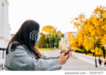 Portrait of a beautiful adult young woman on the background of autumn in the Park in a medical face mask with a phone in her hands Portrait of a beautiful adult young woman on the background of autumn in the Park in a medical face mask with a phone in her hands 70397077
