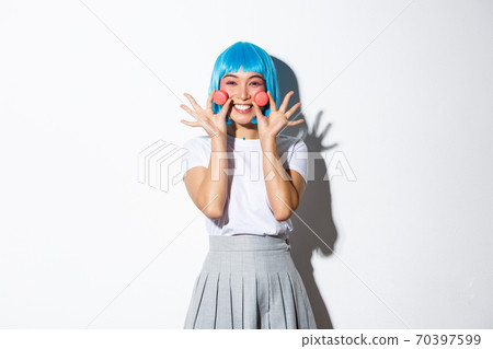 Image of beautiful smiling asian girl looking silly, showing macaroons, wearing blue anime wig, standing over white background 70397599