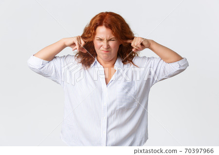 Annoyed and bothered, complaining redhead middle-aged woman grimacing and shut ears with fingers from awful bothering noise, cant stand loud terrible music, standing white background 70397966