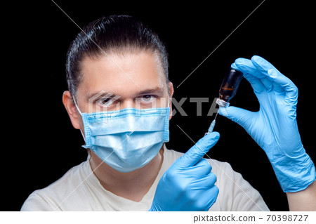 Doctor in a medical mask and sterile blue gloves holds in his hands a syringe with a needle and a glass bottle with a vaccine for viral disease, closeup of a medic isolated on a black background. Doctor in a medical mask and sterile blue gloves holds in his hands a syringe with a needle and a glass bottle with a vaccine for viral disease, closeup of a medic isolated on a black background. 70398727