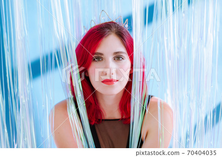Shoulders portrait of attractive red heared woman with vivid make-up and crown on head sit between the shiny stripes of transparent curtain on blue background and look into the camera. Shoulders portrait of attractive red heared woman with vivid make-up and crown on head sit between the shiny stripes of transparent curtain on blue background and look into the camera. 70404035