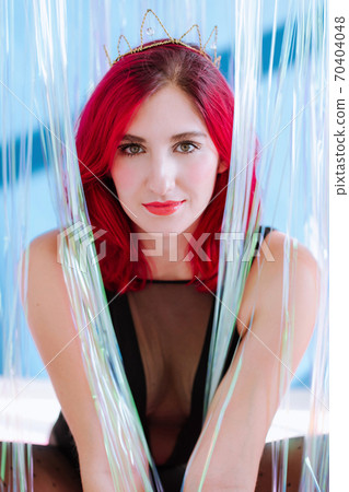 Verticaql crop portrait of beautiful red heared woman with bright makeup sitting between the glitchy stripes of transparent curtain on blue wall and friendly look into camera. 70404048