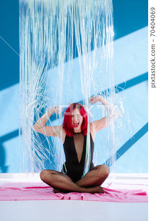 Young girl with red hair sitting on floor in the middle of shiny curtain against blue wall raising her hands loudly scream expressing strong emotion. Respons against reality Young girl with red hair sitting on floor in the middle of shiny curtain against blue wall raising her hands loudly scream expressing strong emotion. Respons against reality 70404069