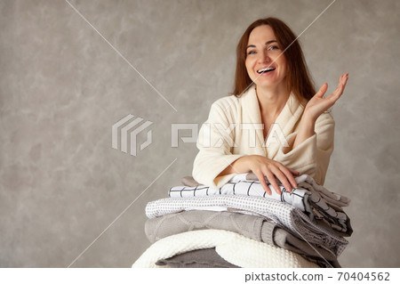 Young woman with beautiful smile in warm beige robe is leaning on stack of folded bed sheets 70404562