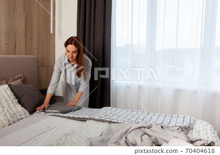 Happy woman with long red hair is folding clean bedding set on the bed of room with copy space. Happy woman with long red hair is folding clean bedding set on the bed of room with copy space. 70404618