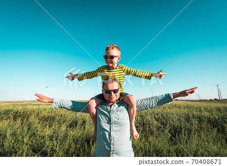 Green renewable energy concept. Happy family playing on field with wind generator turbines. Father carrying little kid boy on shoulders and waving their arms like a windmill. Green renewable energy concept. Happy family playing on field with wind generator turbines. Father carrying little kid boy on shoulders and waving their arms like a windmill. 70408671