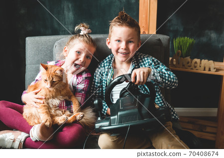 Funny little girl and boy playing game with steering wheel console. Funny kids with pet cat in hands sitting on sofa at home. Happy brother and sister. Family time. Funny little girl and boy playing game with steering wheel console. Funny kids with pet cat in hands sitting on sofa at home. Happy brother and sister. Family time. 70408674