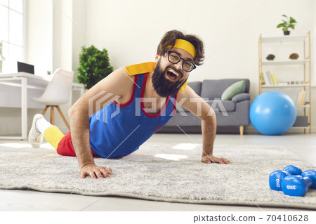 Funny smiling bearded man in colorful sportswear and glasses trains on the floor in the room. 70410628