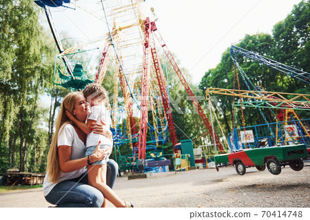 Gives hugs. Cheerful little girl her mother have a good time in the park together near attractions Gives hugs. Cheerful little girl her mother have a good time in the park together near attractions 70414748