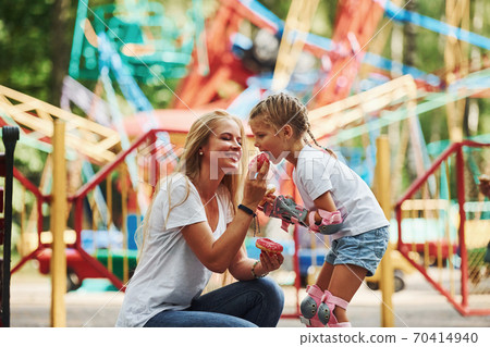 Eating donuts. Cheerful little girl on roller skates and her mother have a good time in the park together near attractions 70414940