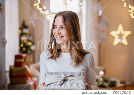 Happy young woman indoors at home at Christmas, holding present. Happy young woman indoors at home at Christmas, holding present. 70417668
