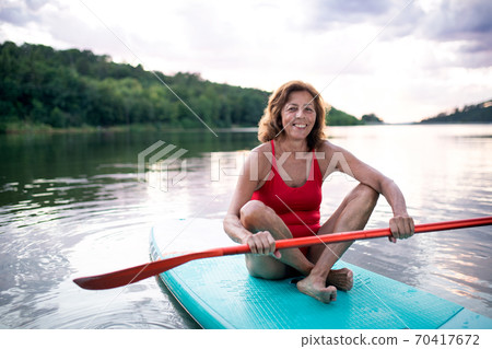 Senior woman paddleboarding on lake in summer. Copy space. 70417672