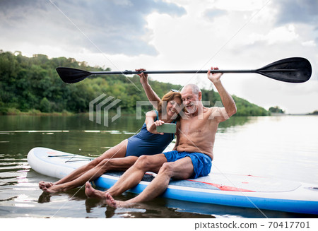 Senior couple sitting on paddleboard on lake in summer, taking selfie with smartphone. Senior couple sitting on paddleboard on lake in summer, taking selfie with smartphone. 70417701