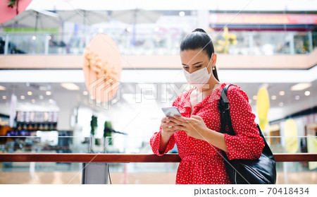 Woman with face mask standing with smartphone indoors in shopping center, coronavirus concept. Woman with face mask standing with smartphone indoors in shopping center, coronavirus concept. 70418434