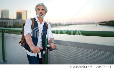 Mature man commuter with electric scooter outdoors in city, going to work. Copy space. Mature man commuter with electric scooter outdoors in city, going to work. Copy space. 70418481