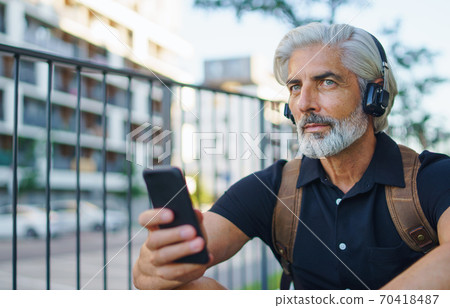 Portrait of mature man with headphones sitting outdoors in city, using smartphone. Portrait of mature man with headphones sitting outdoors in city, using smartphone. 70418487