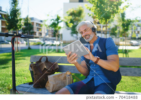 Portrait of mature man with headphones sitting outdoors in city, using tablet. Portrait of mature man with headphones sitting outdoors in city, using tablet. 70418488
