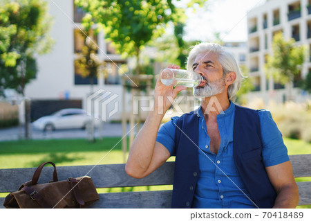 Portrait of mature man sitting outdoors in city, drinking water. Portrait of mature man sitting outdoors in city, drinking water. 70418489