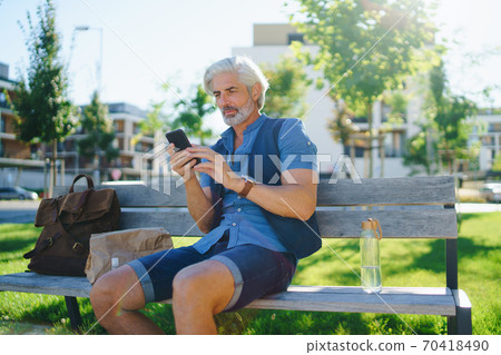 Portrait of mature man sitting outdoors on bench in city, using smartphone. Portrait of mature man sitting outdoors on bench in city, using smartphone. 70418490