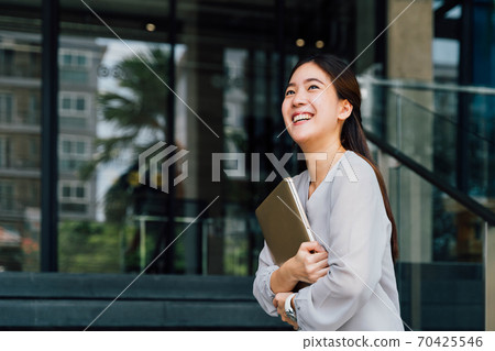 One successful and happy young adult business woman holding a laptop with smile. Professional female worker standing in corporate modern office and window in casual gray look. - With copy space One successful and happy young adult business woman holding a laptop with smile. Professional female worker standing in corporate modern office and window in casual gray look. - With copy space 70425546