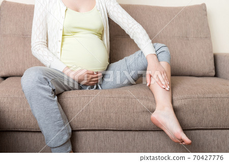 Leg cramps during pregnancy. Closeup of hands massaging swollen foot while sitting on sofa 70427776