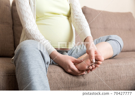 Leg cramps during pregnancy. Closeup of hands massaging swollen foot while sitting on sofa 70427777