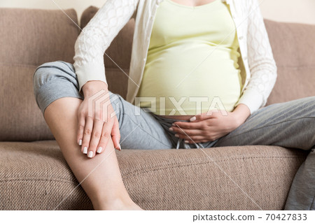 Leg cramps during pregnancy. Closeup of hands massaging swollen foot while sitting on sofa 70427833