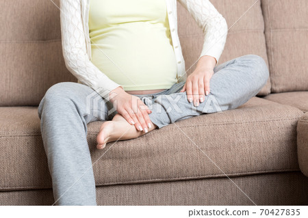 Leg cramps during pregnancy. Closeup of hands massaging swollen foot while sitting on sofa 70427835