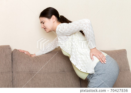 beautiful pregnant woman touching her tummy and keeping one hand on her back at home on sofa 70427836