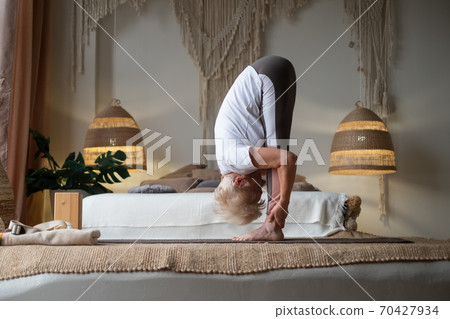 Attractive senior woman working out standing in Uttanasana at home. Attractive senior woman working out standing in Uttanasana at home. 70427934