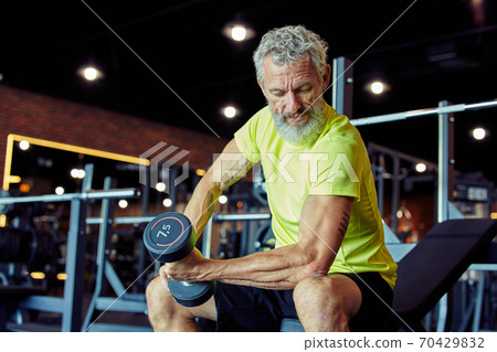 Weight workout. Focused mature man in sportswear lifting heavy dumbbells and pumping his biceps while training at gym Weight workout. Focused mature man in sportswear lifting heavy dumbbells and pumping his biceps while training at gym 70429832