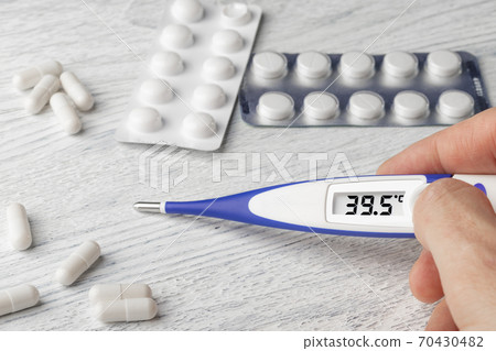 White-blue thermometer with a high temperature of 39.5 degrees Celsius in hand with pills and medical capsules on a wooden background 70430482