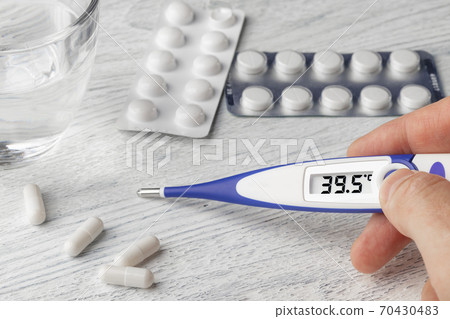 White-blue thermometer with a high temperature of 39.5 degrees Celsius in hand with pills and medical capsules on a wooden background 70430483