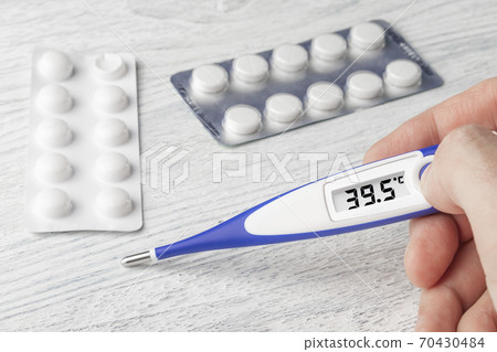 White-blue thermometer with a high temperature of 39.5 degrees Celsius in hand with pills and medical capsules on a wooden background 70430484