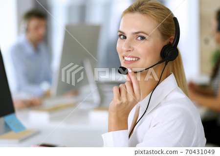 Blonde business woman using headset for communication and consulting people at customer service office. Call center. Group of operators at work at the background Blonde business woman using headset for communication and consulting people at customer service office. Call center. Group of operators at work at the background 70431049