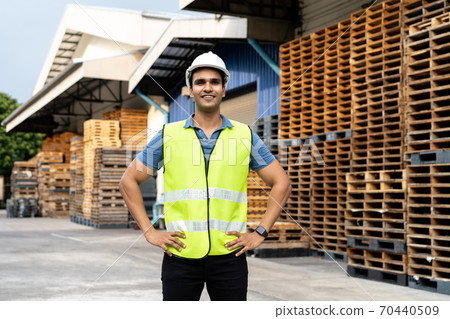 Portrait of young Indian worker working in logistic industry outdoor in front of factory warehouse. Smiling happy man with hard hat looking at camera hands on hip at depot Portrait of young Indian worker working in logistic industry outdoor in front of factory warehouse. Smiling happy man with hard hat looking at camera hands on hip at depot 70440509