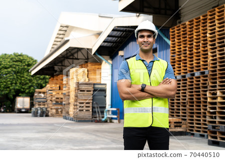 Portrait of young Indian worker with arm folded working in logistic industry in front of factory warehouse. Smiling happy man in hard hat looking at camera arms crossed at depot 70440510