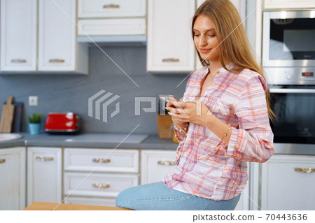 Pretty young woman in casual clothes using her smartphone in kitchen 70443636