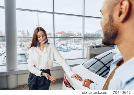 Young attractive woman salesperson in car showroom showing a car to her male client Young attractive woman salesperson in car showroom showing a car to her male client 70443642