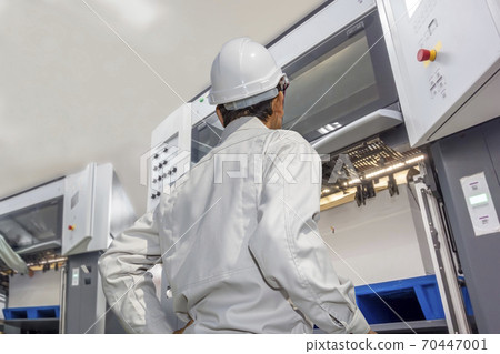 The upper body of the back of a male operator wearing work clothes operating a sheet-fed offset printing machine at a printing factory. 70447001