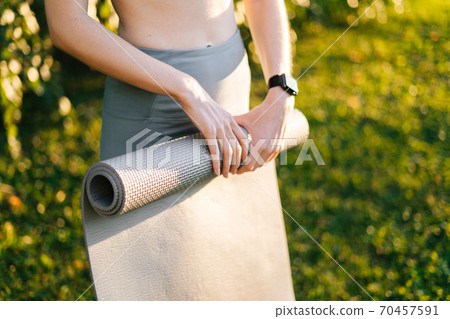 Close-up of hands of unrecognizable young woman rolling yoga mat after exercising at city park. 70457591