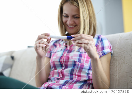 Happy woman is sitting on couch and looking at pregnancy test Happy woman is sitting on couch and looking at pregnancy test 70457858