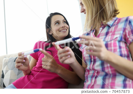Two women are sitting on couch holding two pregnancy tests. Two women are sitting on couch holding two pregnancy tests. 70457873