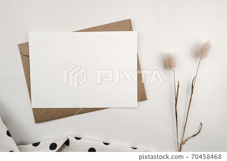 Blank white paper on brown paper envelope with Rabbit tail dry flower and White cloth with black dots. Mock-up of horizontal blank greeting card. Top view of Craft envelope on white background. 70458468