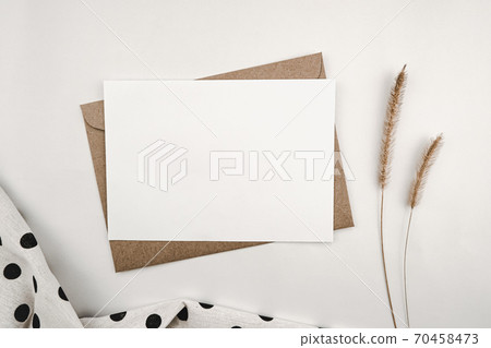 Blank white paper on brown paper envelope with Bristly foxtail dry flower and White cloth with black dots. Mock-up of horizontal blank greeting card. Top view of Craft envelope on white background. 70458473
