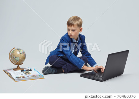 blond boy in a school suit is sitting in front of a laptop, a globe, and a book. photo session in the Studio on a white background blond boy in a school suit is sitting in front of a laptop, a globe, and a book. photo session in the Studio on a white background 70460376