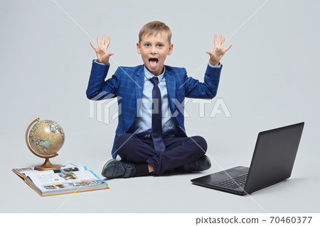 blond boy in a school suit is sitting in front of a laptop, a globe, and a book. photo session in the Studio on a white background blond boy in a school suit is sitting in front of a laptop, a globe, and a book. photo session in the Studio on a white background 70460377