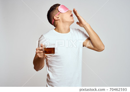 handsome man with pink sleep mask and with a cup of tea in hand pulls himself up on a light background handsome man with pink sleep mask and with a cup of tea in hand pulls himself up on a light background 70462593